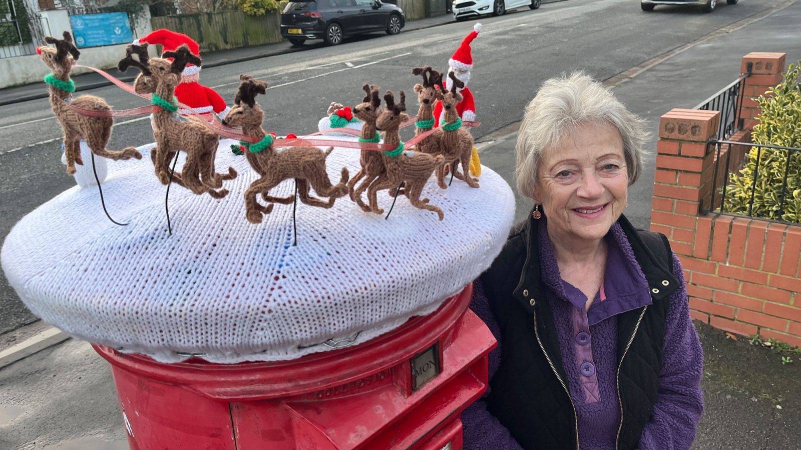 A woman with grey hair stands next to a postbox with an elaborate knitted topper including santa's sleigh and reindeer