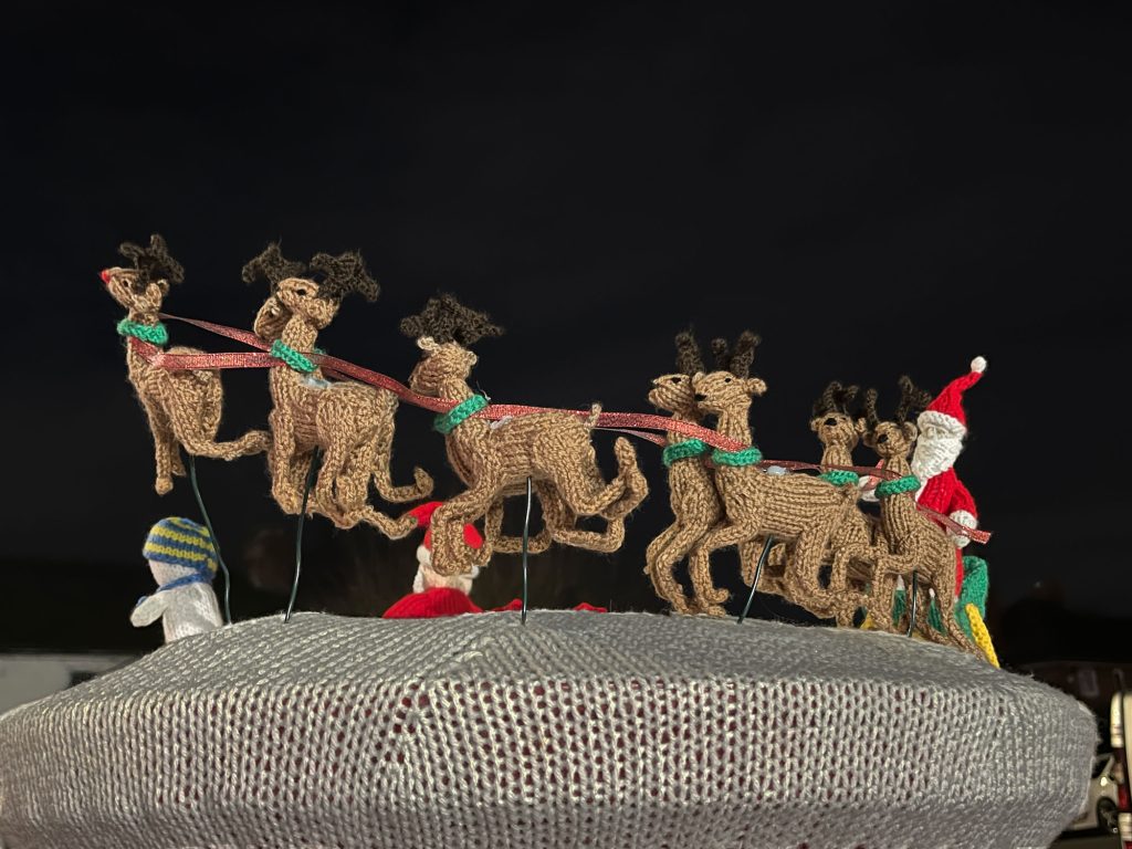 A knitted postbox topper of santa's sleigh pulled by reindeer, with Rudolph at the front