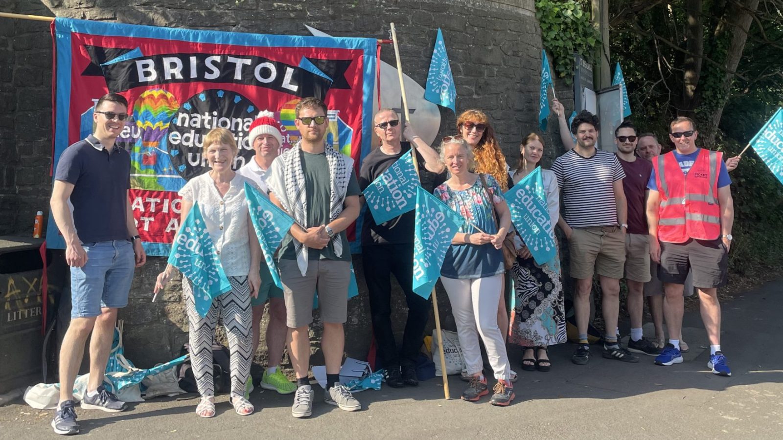 A group of teachers holding blue National Education Union flags stand in front of a brick wall