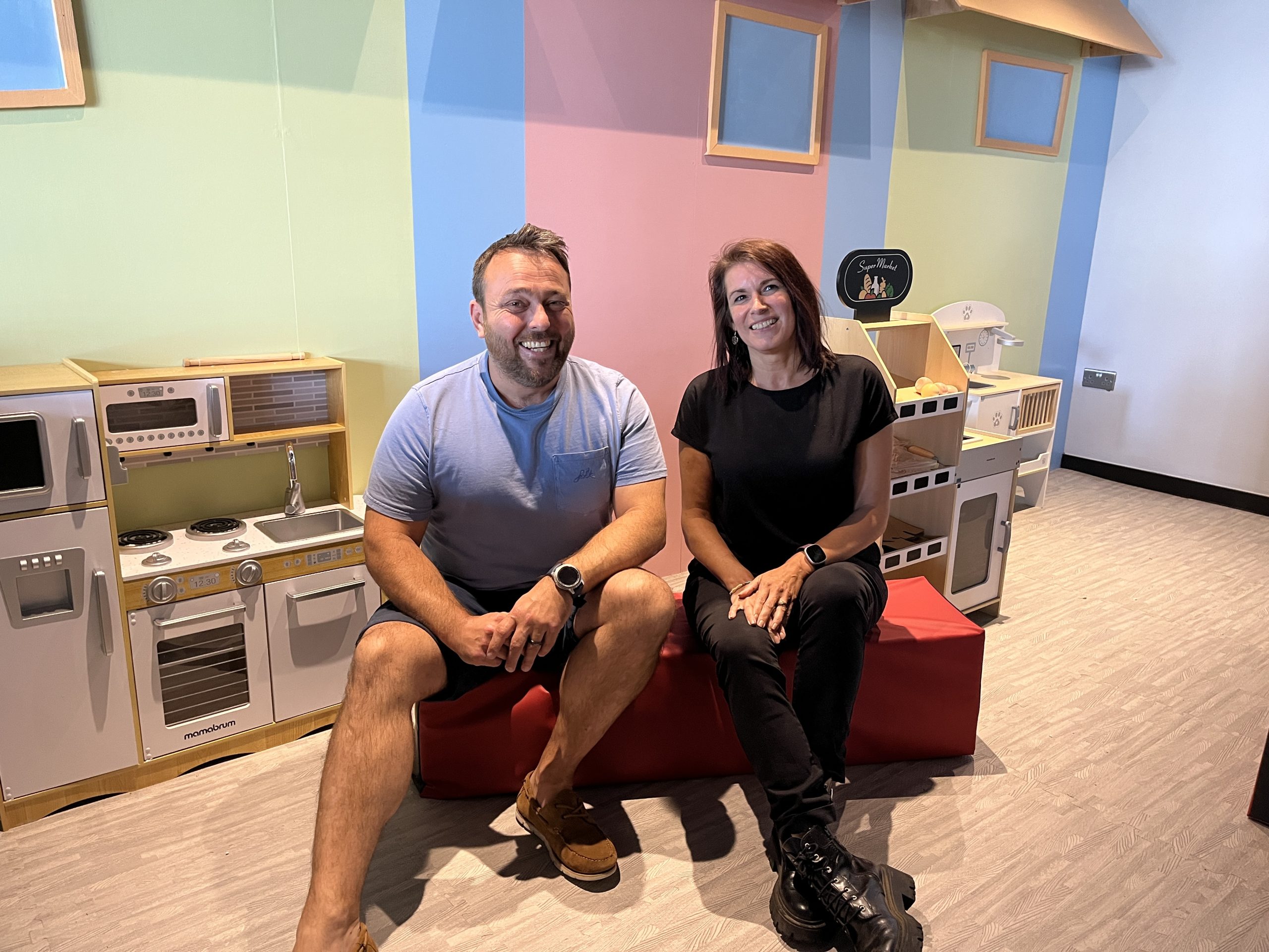 A man and a women sit in front of some miniture kitchen appliances for childrens play