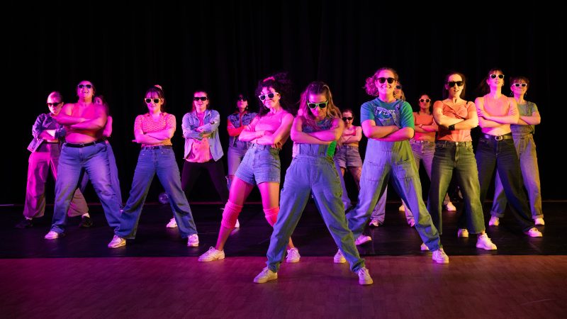 A group of dancers wearing dungarees, trainers and sunglasses, posing with their arms crossed