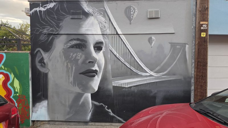 Monochrome mural of a woman from the neck up. Her hair is tied back and 1958 is written on her cheek. The mural continues to her right with a painting of Clifton Suspension Bridge