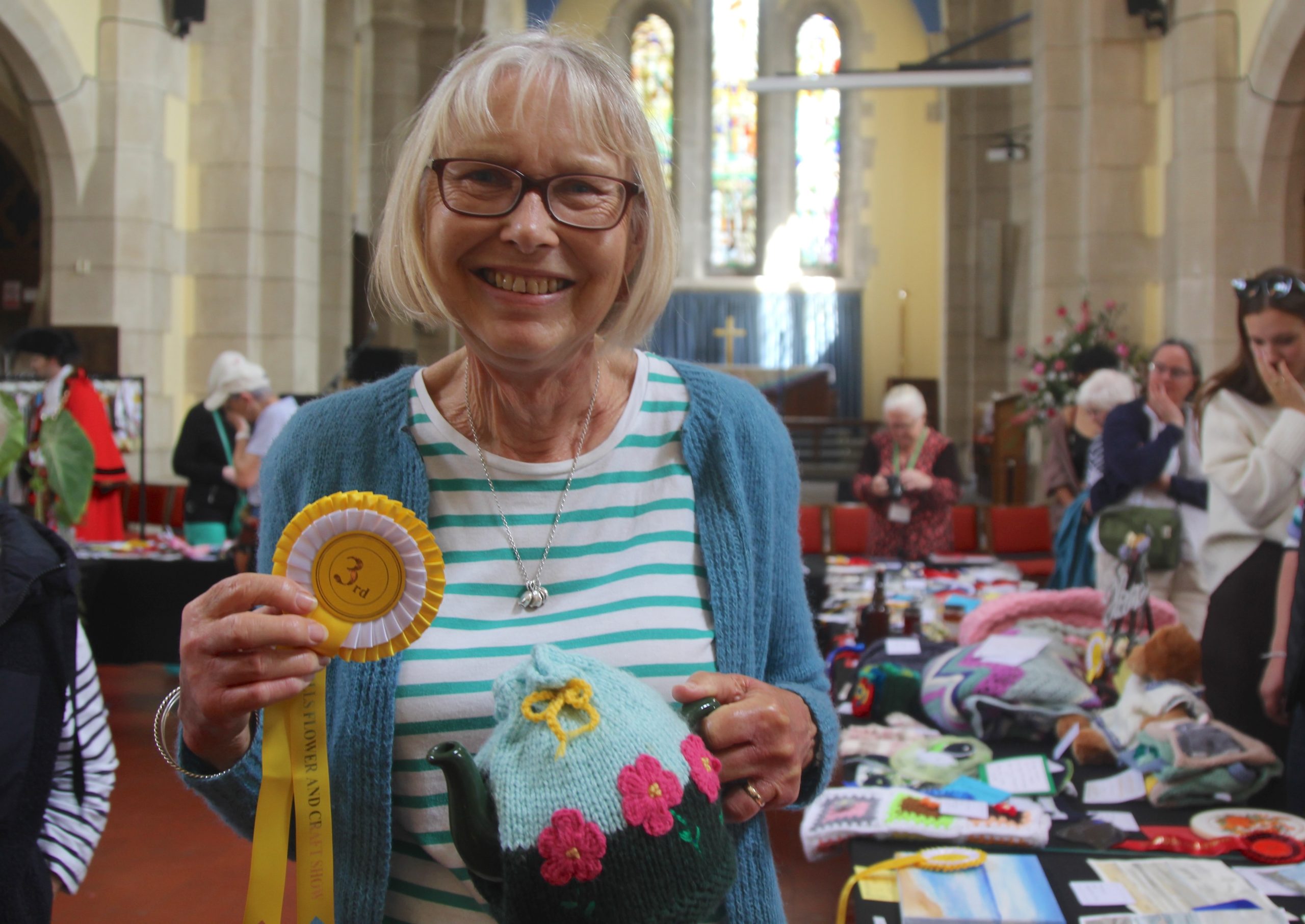 A smiling woman holds a rosette and a teapot covered in a knitted, flowered tea cosy