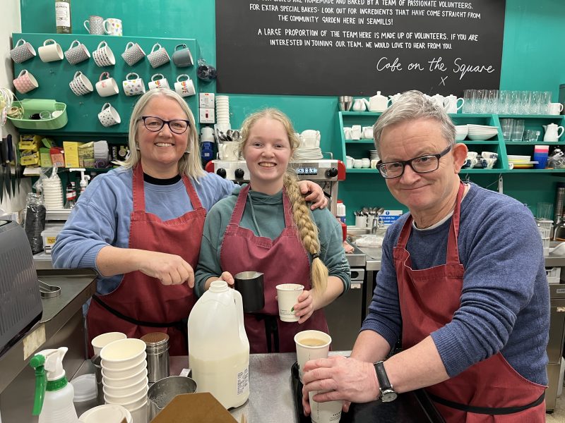 Three smiling people prepare hot drinks at a cafe counter