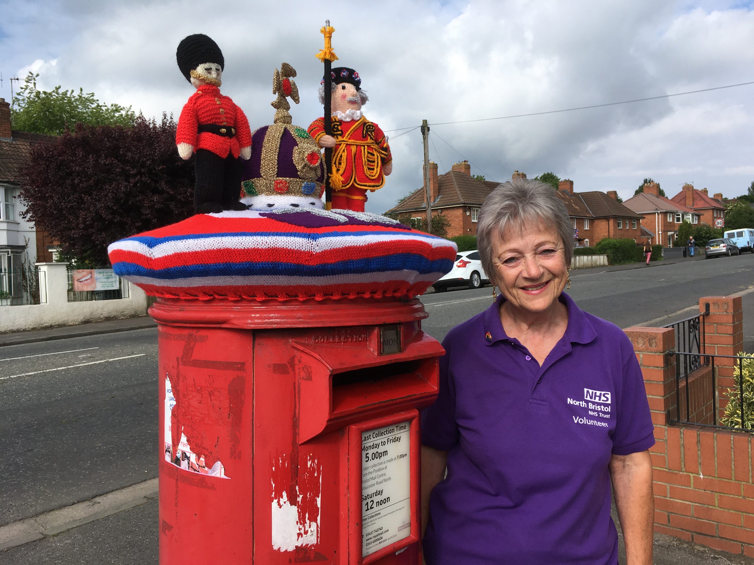 A woman stands next to a postbox with a knitted topper featuring a beefeater and a crown on it